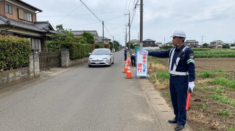 民家と畑が隣接する野田市の狭路工事現場で見通し確認と車両誘導を行う警備員